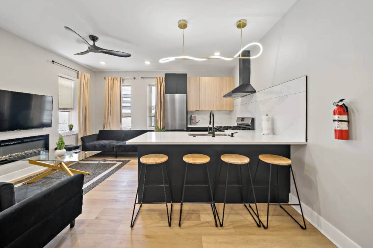 Modern kitchen featuring brand-new appliances, including a breakfast bar, dishwasher, microwave, and refrigerator. It seamlessly connects to the living room, which is furnished with 2 sofa beds, fireplace ,and a flat-screen TV.