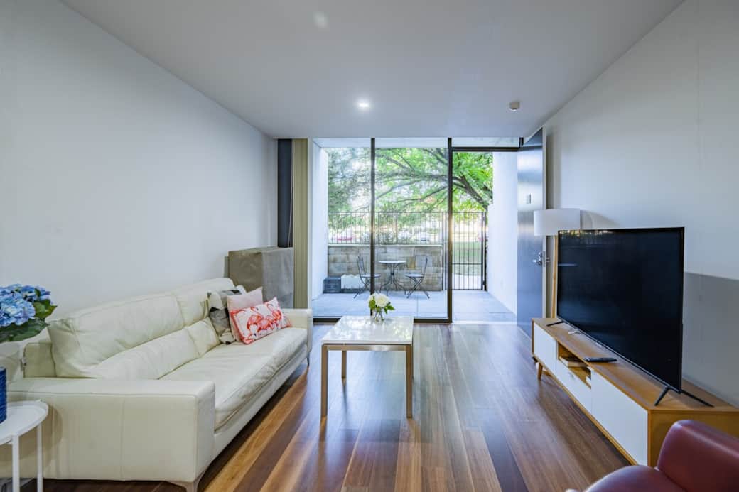Bask in the natural light that floods our contemporary living room. The minimalist design features a white sofa, a sleek coffee table, and a TV, creating a tranquil environment for relaxation.