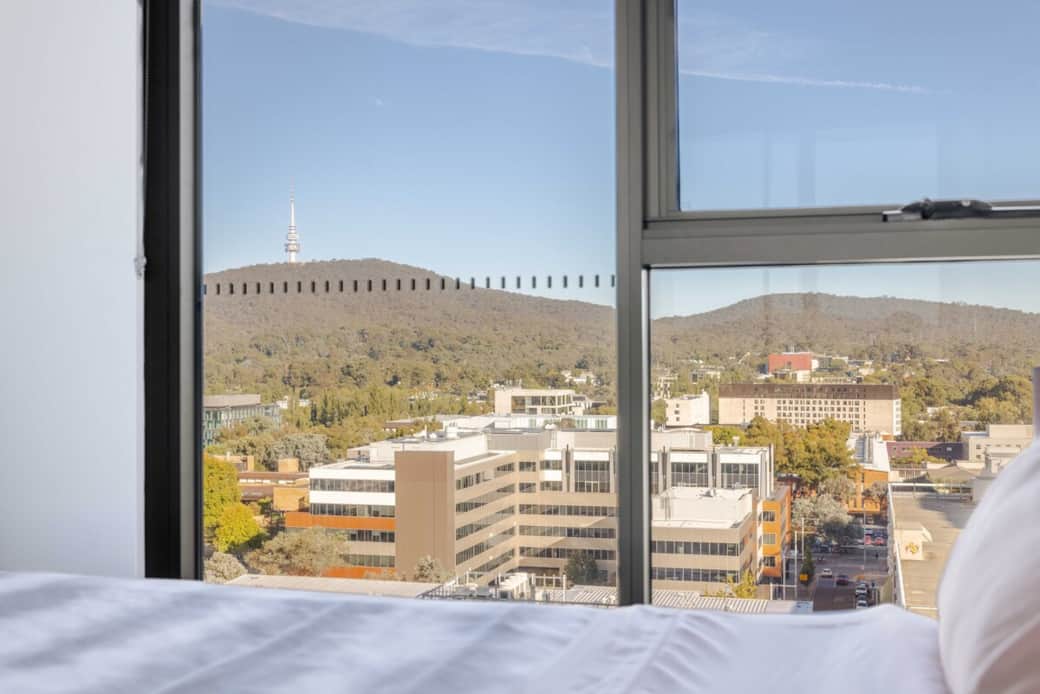 Wake up to stunning city views from the comfort of your bed. This bedroom features a large window that frames the urban landscape and hills beyond, offering a serene backdrop to your stay. The white linens and cozy ambiance make it a perfect retreat.