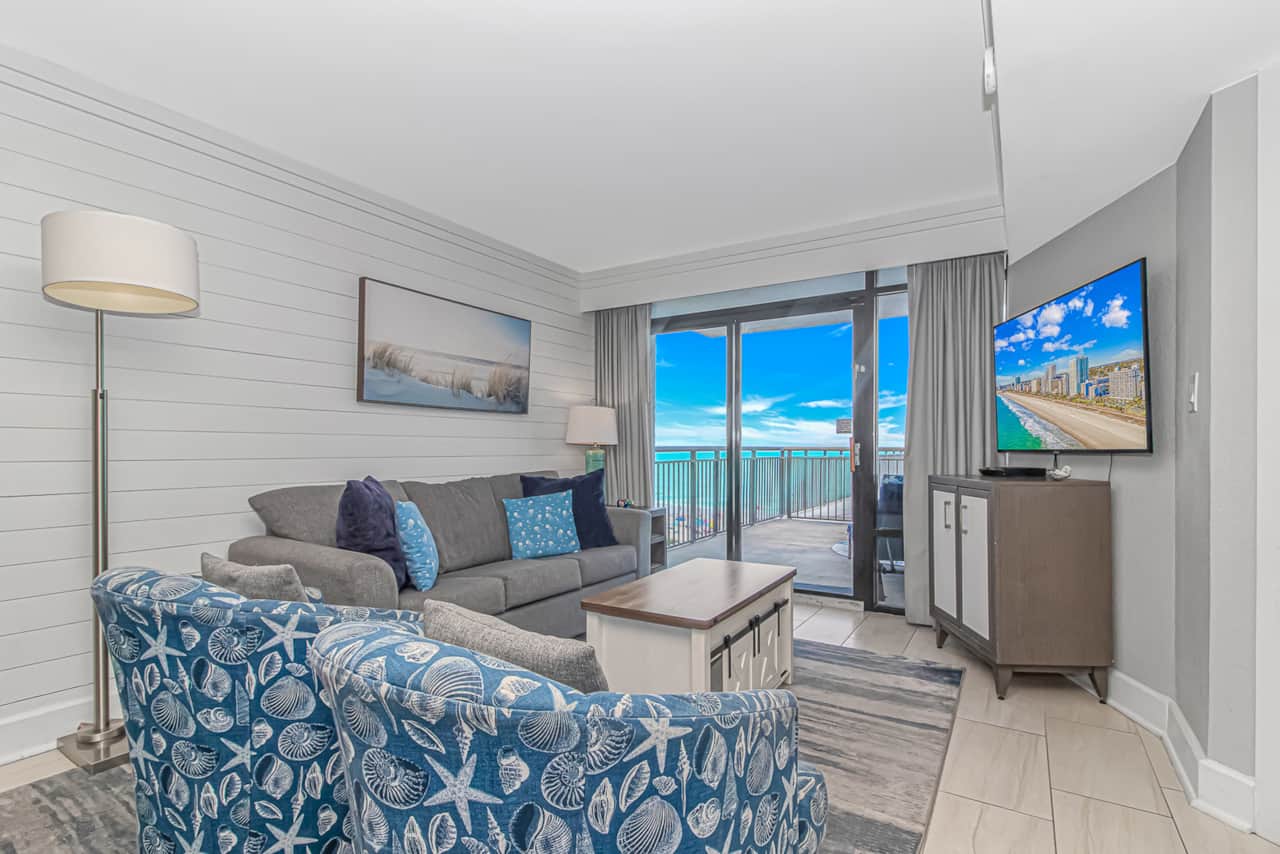 Bright coastal-inspired living room featuring comfortable seating, Smart TV, and floor-to-ceiling balcony access with breathtaking oceanfront views.