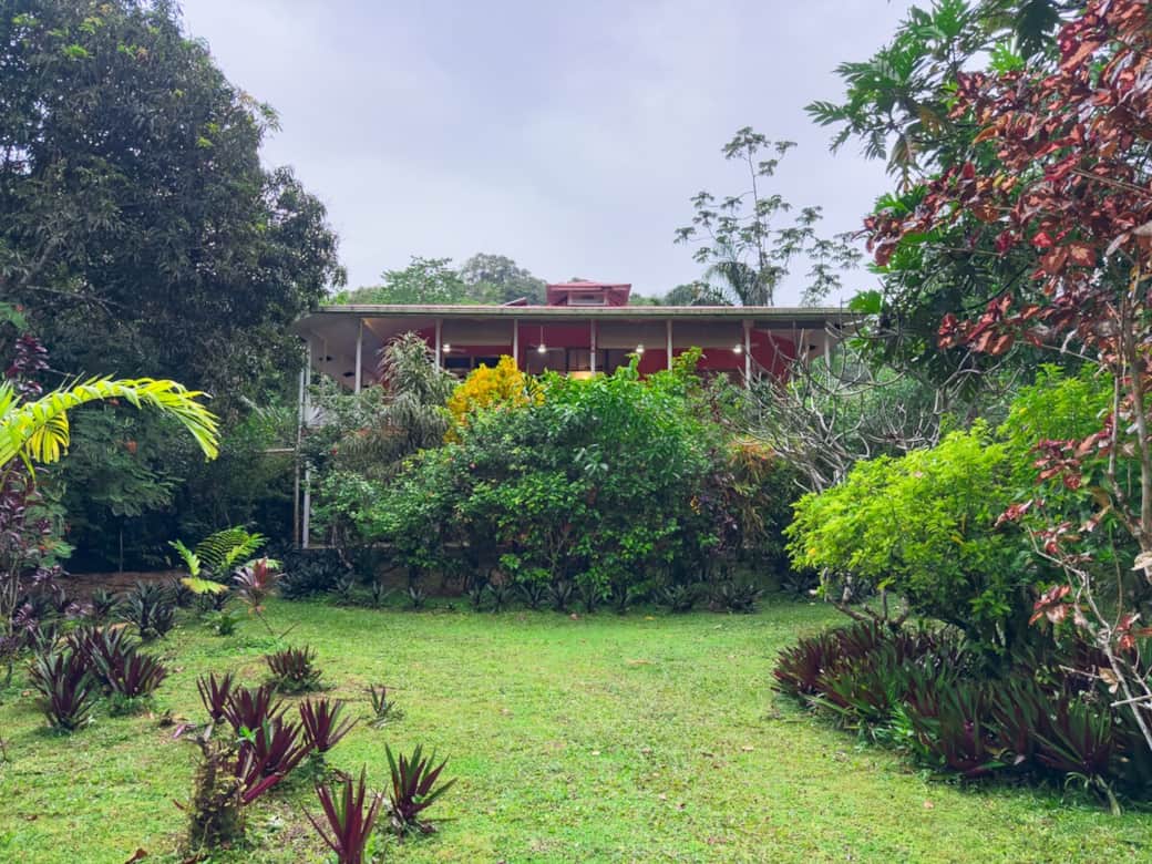 Front façade featuring covered upstairs terrace, ceiling fans, and warm lighting above the lush garden — ideal for indoor-outdoor Caribbean living.
