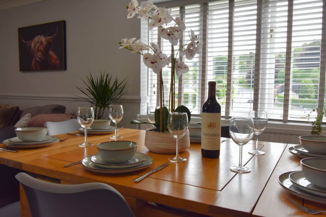 A bright, welcoming dining area featuring a modern wooden table set for six with stylish crockery and glassware. Large windows offer leafy views, while soft furnishings and decor create a relaxed, elegant space to dine and unwind.