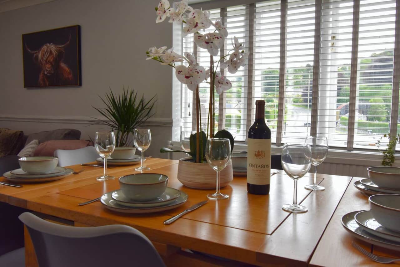 A bright, welcoming dining area featuring a modern wooden table set for six with stylish crockery and glassware. Large windows offer leafy views, while soft furnishings and decor create a relaxed, elegant space to dine and unwind.