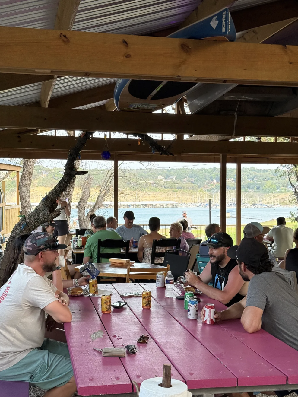 A group of people sitting at pink picnic tables in an outdoor dining area with a lake view.