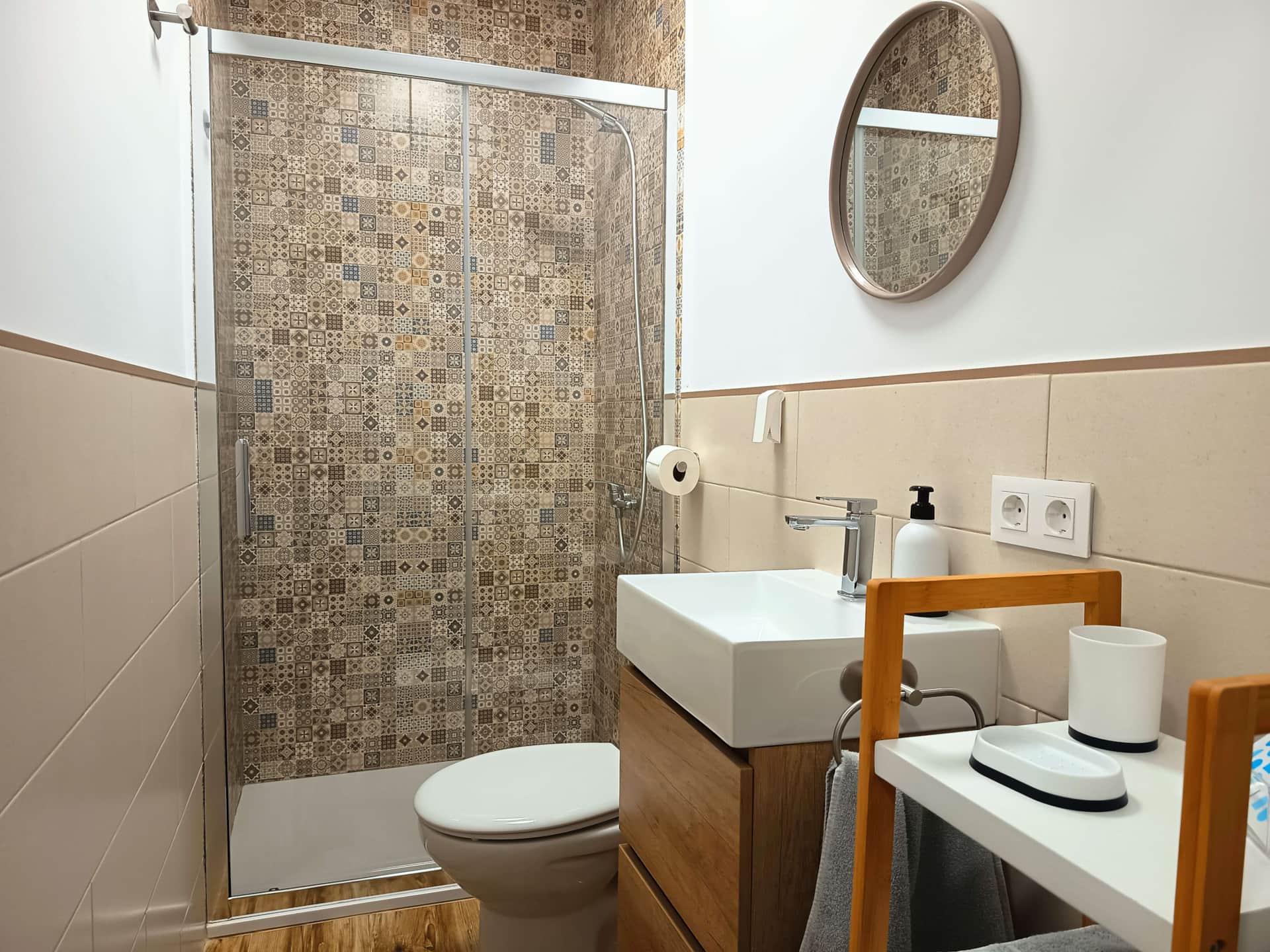 Baño moderno con ducha de mampara de cristal, azulejos decorativos, lavabo con mueble de madera y espejo redondo