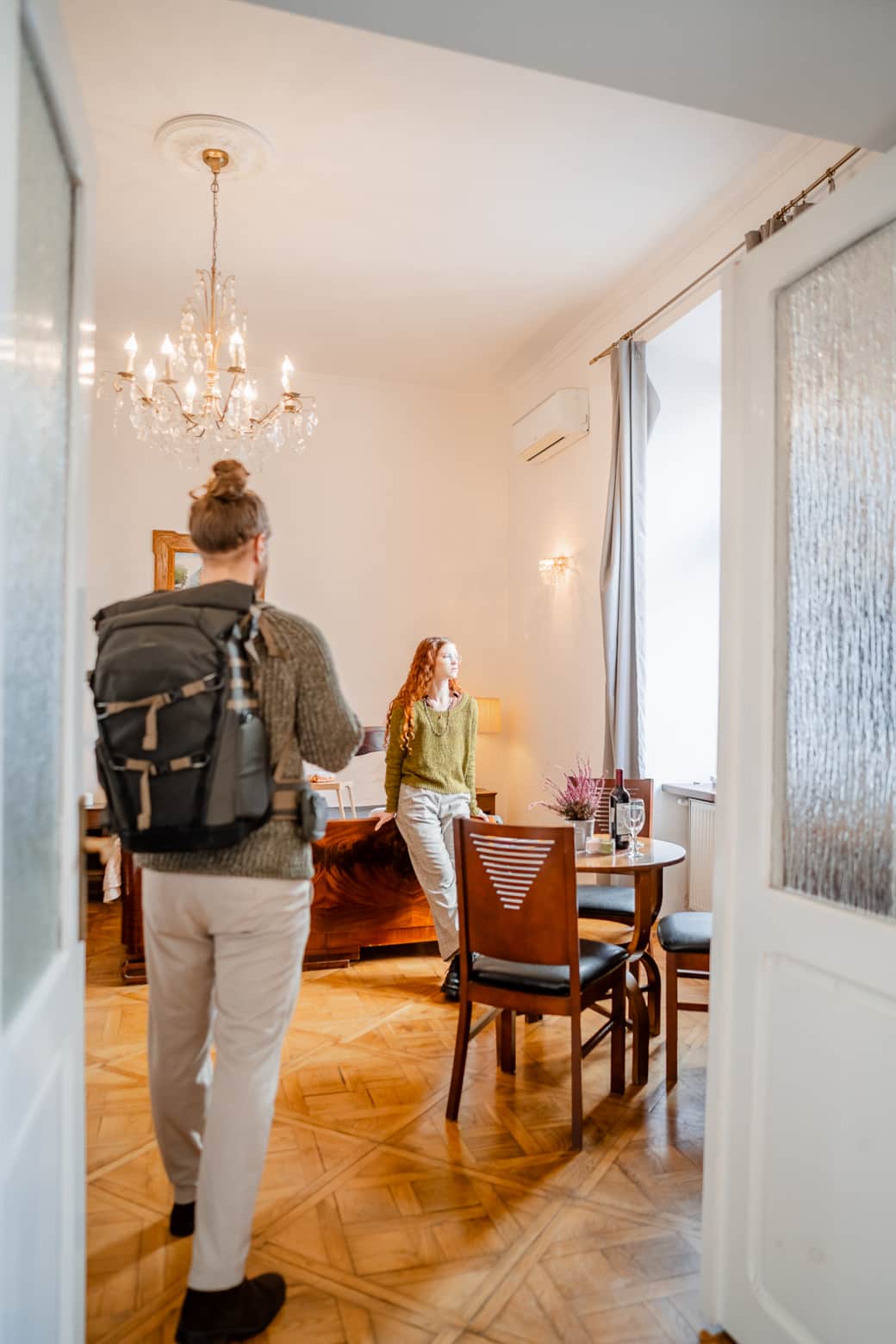 A bright, spacious room with an elegant chandelier, wooden flooring, and a large window.