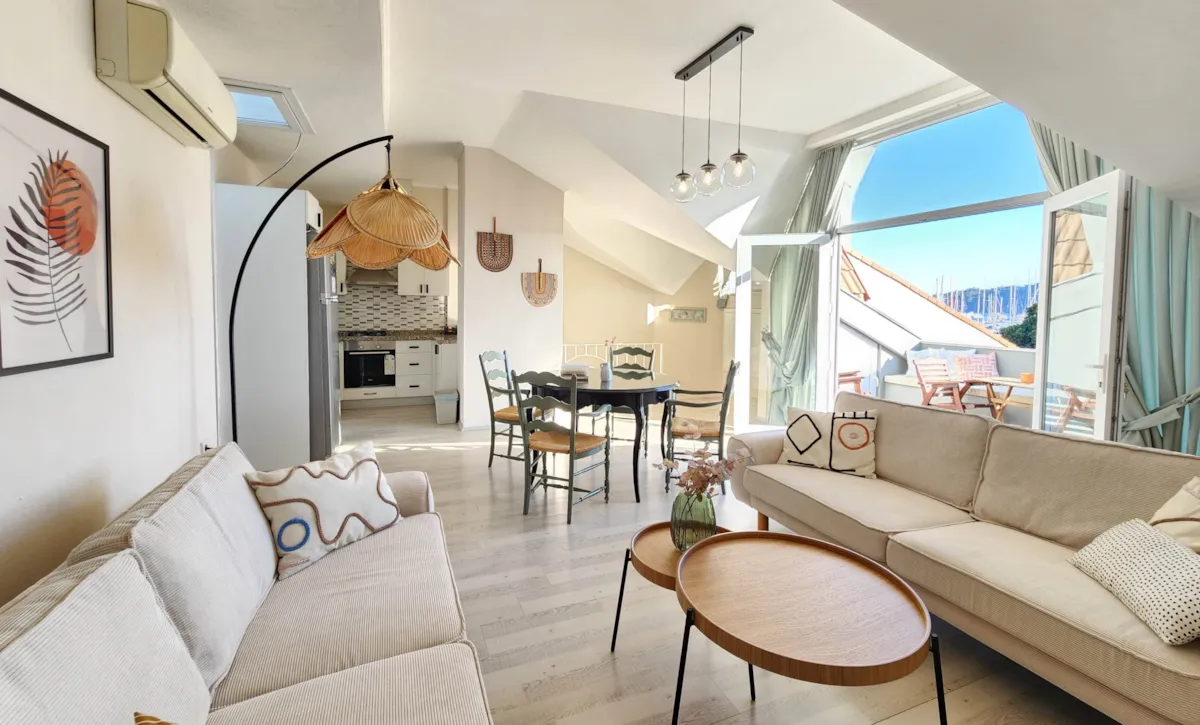 Living area with a sofa, dining table, and kitchen, featuring large windows and a view of the outdoors.