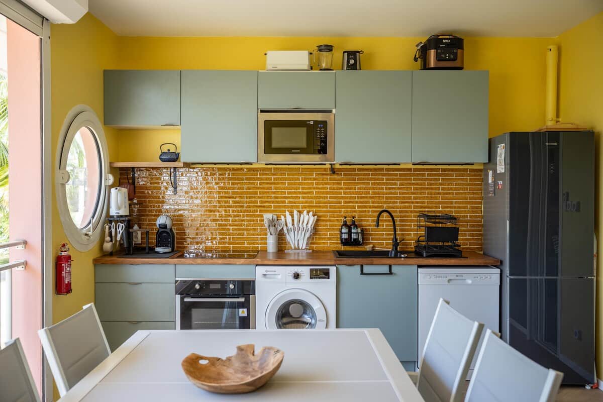 Cuisine équipée avec mur jaune, armoires grises, machine à laver et briques dorées