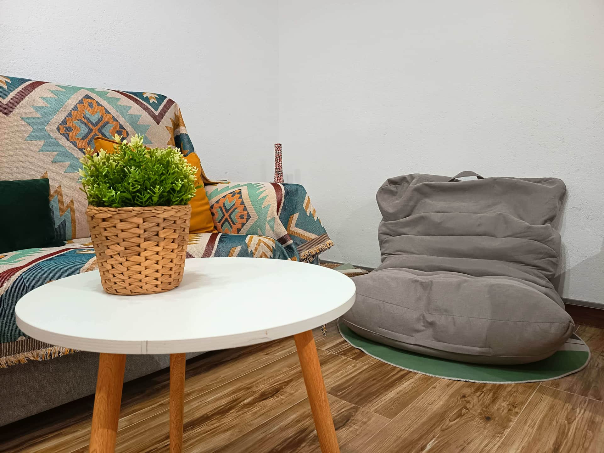 Rincón del salón con sofá decorado con manta de colores, cojín verde, mesa de centro blanca y puff gris sobre alfombra verde