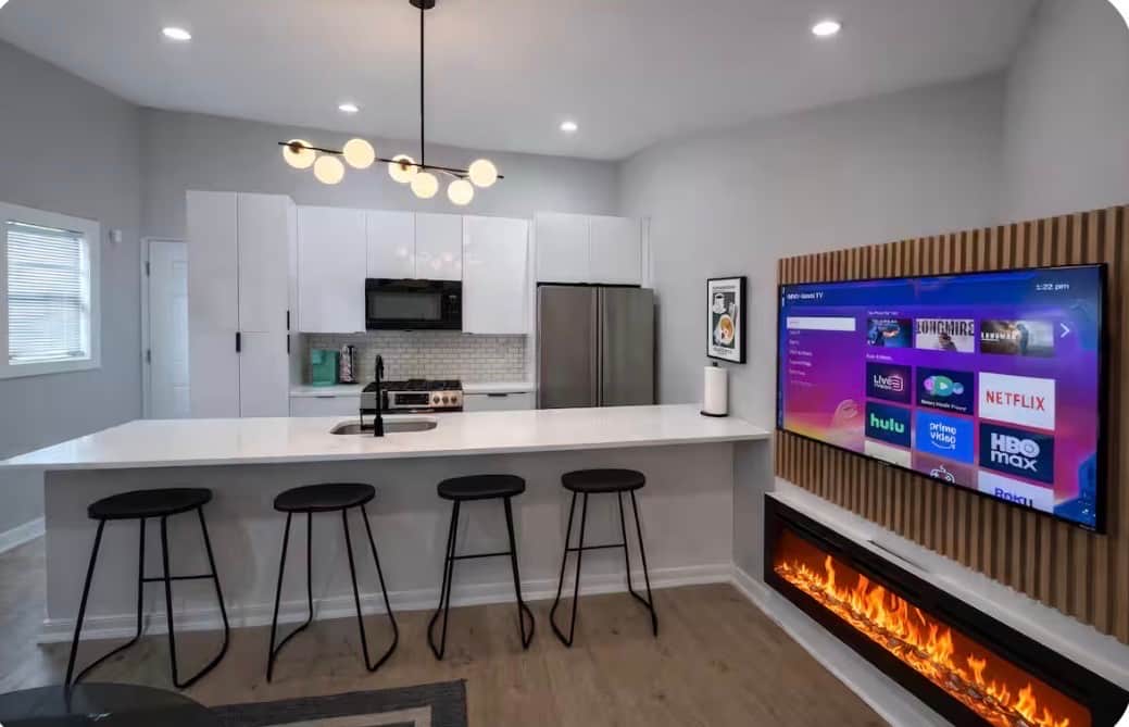Stylish  living space featuring two futon sofa beds, a flat-screen TV, and a modern electric fireplace. The large island bar offers generous seating and flows into the kitchen, creating a comfortable space to relax, dine, or entertain.