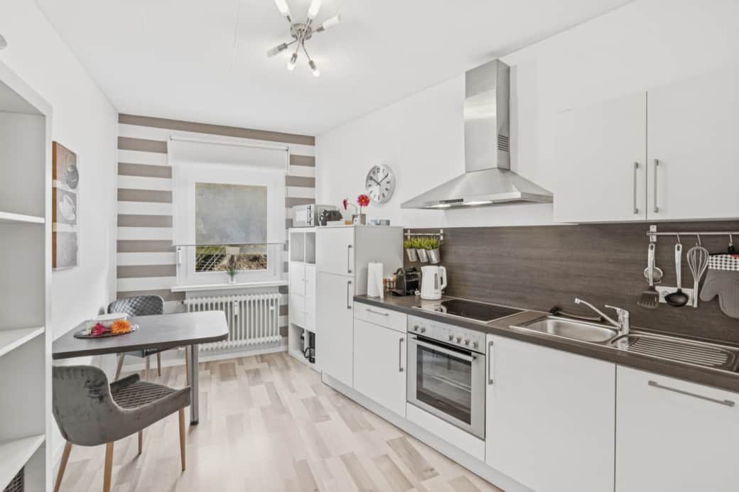 Weiße L-Küche mit grauer Arbeitsplatte, Dunstabzugshaube, Backofen und hellem Holzparkett.