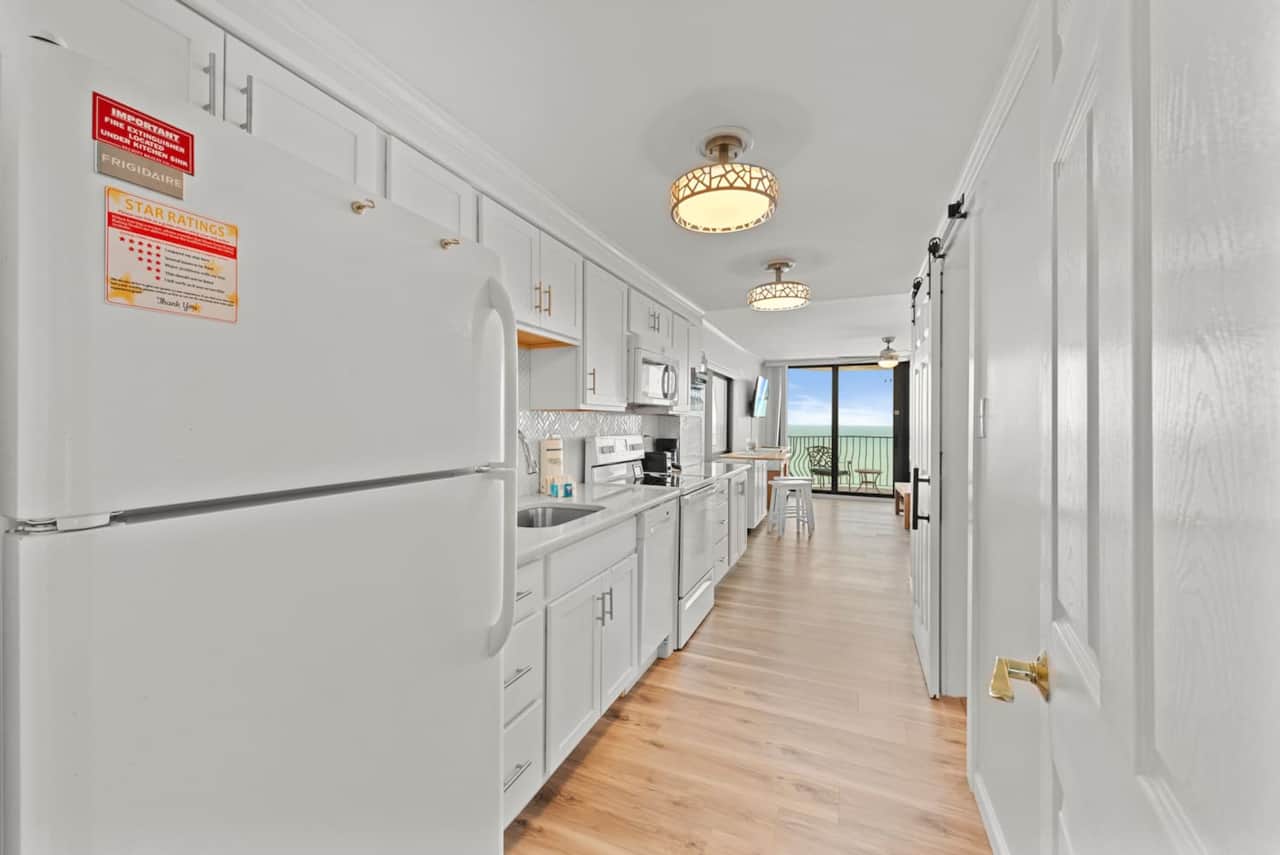 Sleek, modern galley kitchen with bright white cabinetry, updated appliances, and a direct view to the oceanfront balcony — perfect for easy coastal cooking. ️