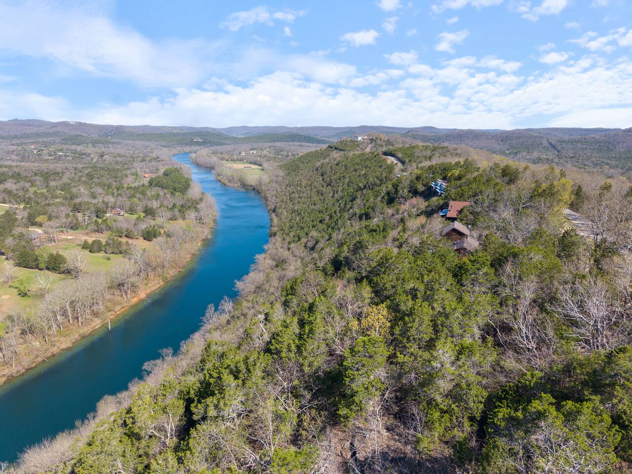 An aerial view of a river running through a wooded area with buildings and hills in the background.