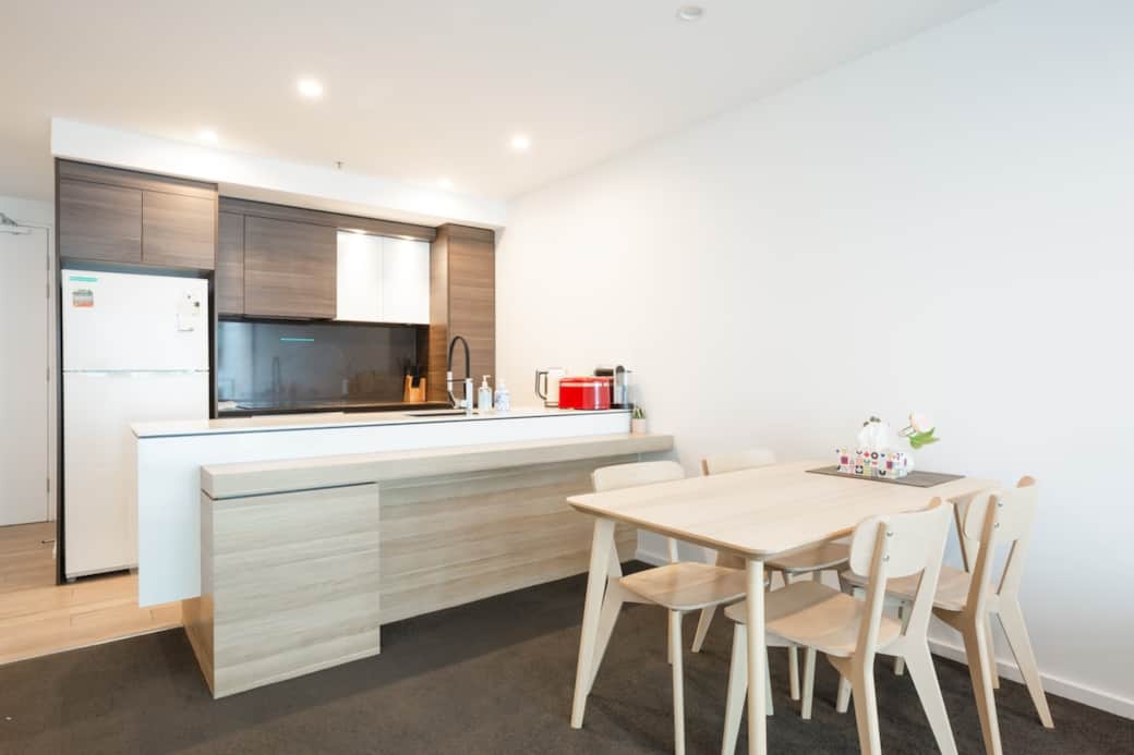 The dining area is set against a backdrop of large windows that offer a view of  outdoors, making meals a delightful experience. The dining table and chairs provide a clean and elegant setting. A vase with flowers adds a touch of nature to the space