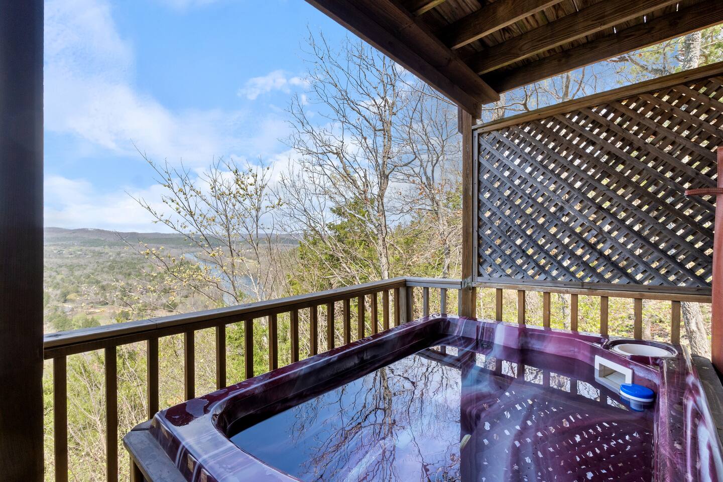 A purple hot tub on a wooden deck with a railing and latticed wall, overlooking a scenic view of trees and a blue sky.