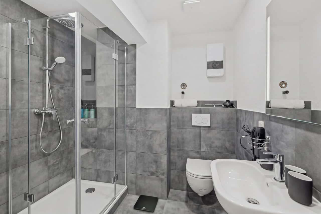 Graue Fliesendusche mit Glastür, Wandhänge-WC, klare Linien, anthrazitfarbene Fliesen.