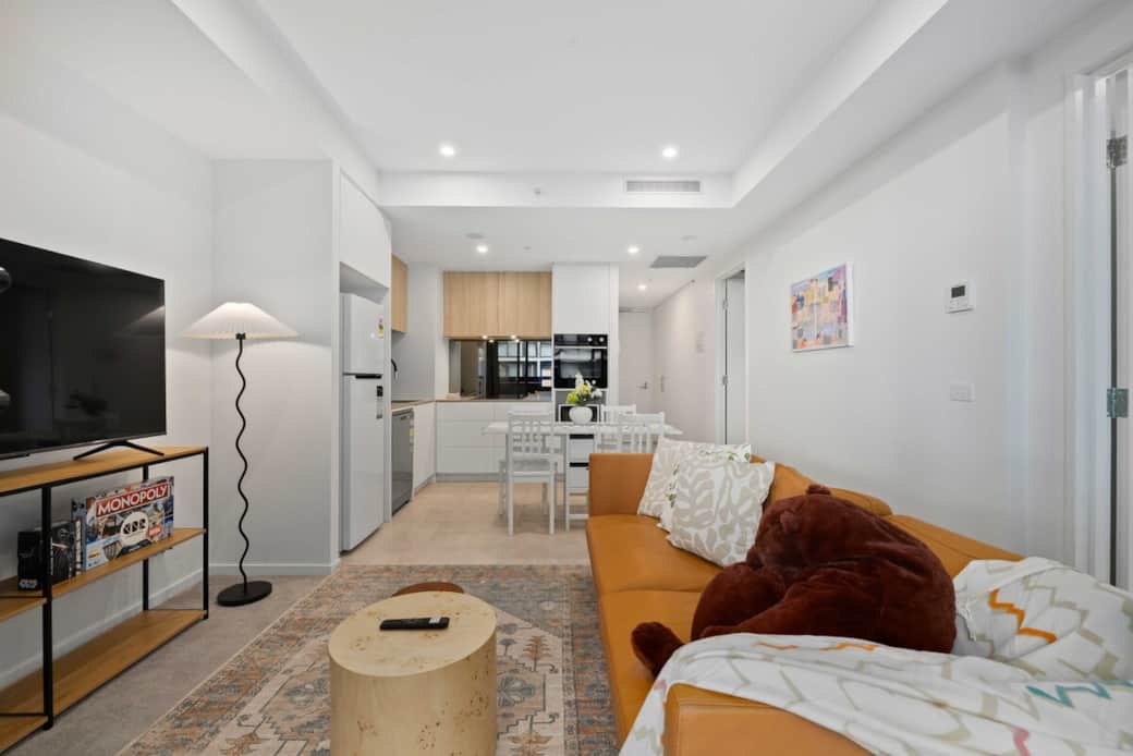 This living room is a perfect blend of comfort and style. The vibrant mustard-yellow sofa, accented with plush pillows, offers a cozy spot to relax. The open layout seamlessly connects to the kitchen, creating an ideal space for both entertainment.
