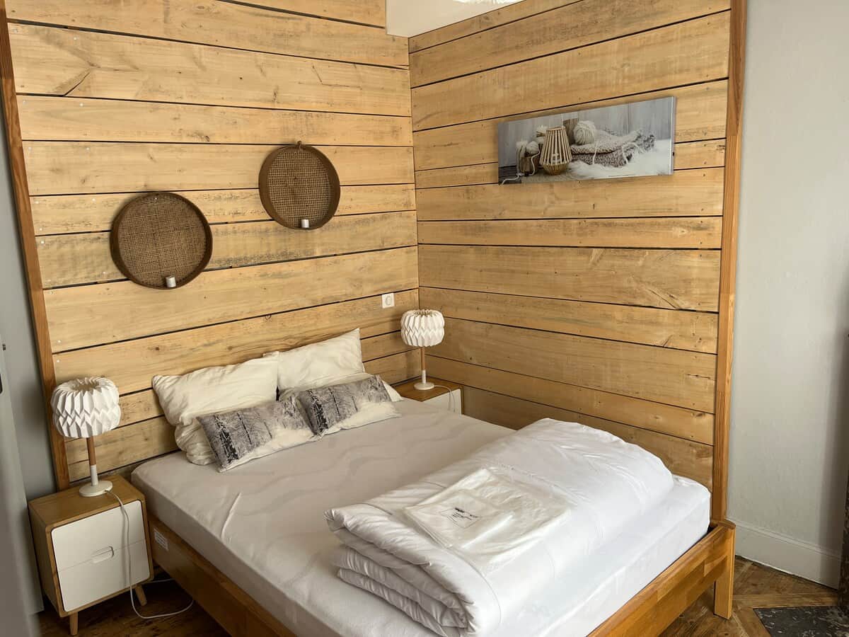 Chambre cosy avec lit double en bois, murs en lambris clair, tables de chevet et lampes design. Décor sobre avec paniers muraux et tableau aux tons naturels, ambiance chaleureuse et apaisante.