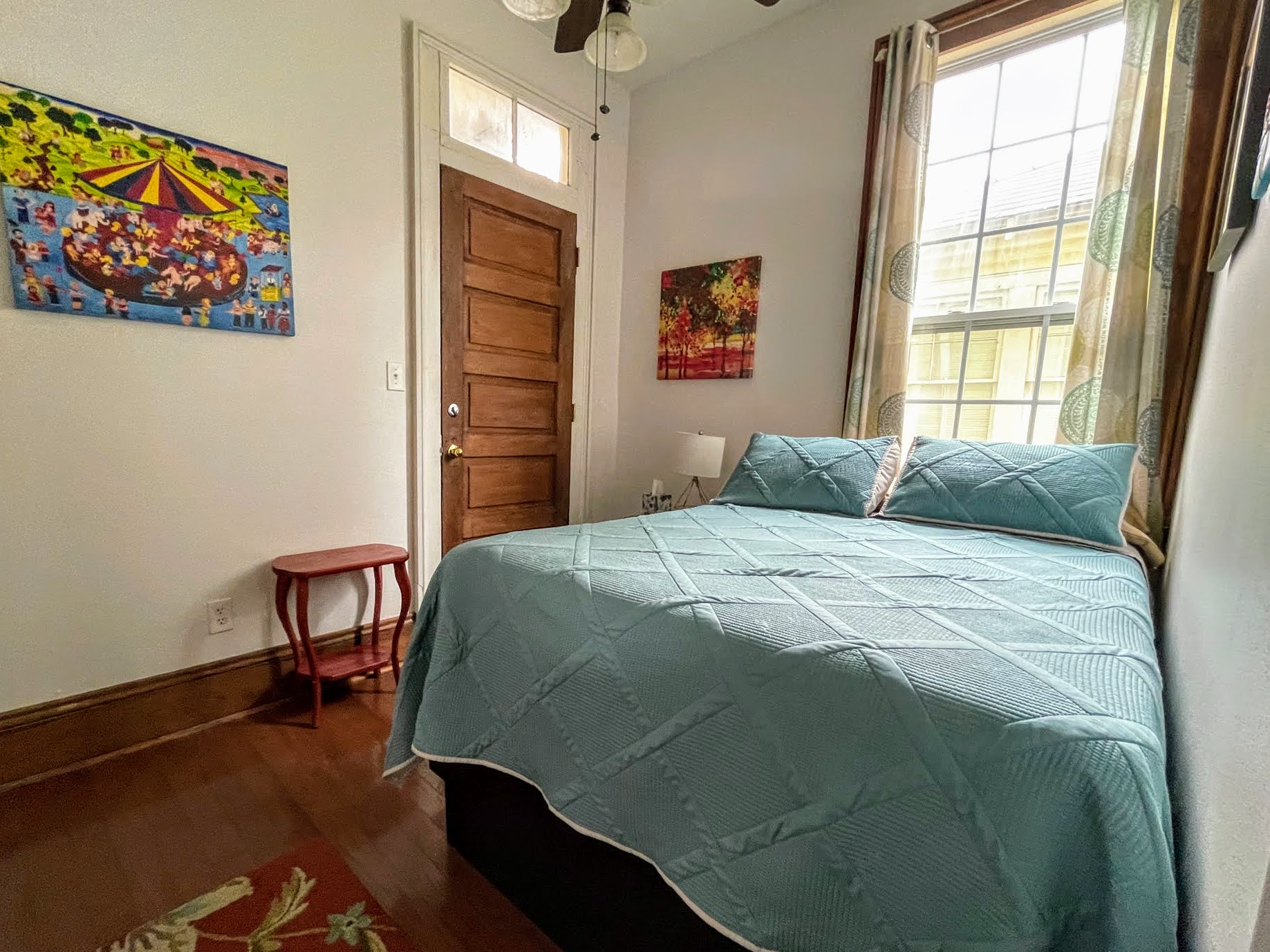 A neatly made bed with teal bedding in a room with white walls, a wooden door, and a window with curtains.
