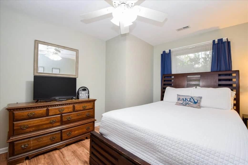 Sun-drenched queen guest room with honey-wood bed, navy curtains, and extra sleeper seating. Queen bed, lake-themed decor, flat-screen TV, gray walls, coastal accent pillow, driftwood flooring, Smith Mountain Lake getaway.