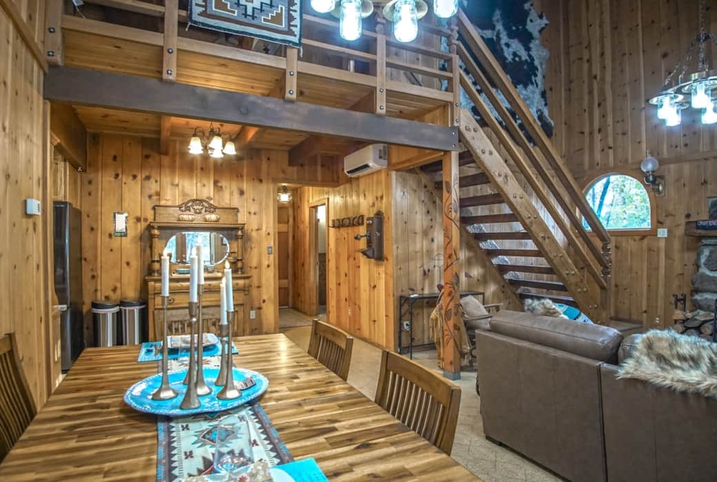 Open-concept cabin living with soaring wood ceilings, a dramatic staircase and loft, and a rustic-elegant dining table styled with turquoise accents—finished with remote-controlled flameless candles for an easy, cozy glow.