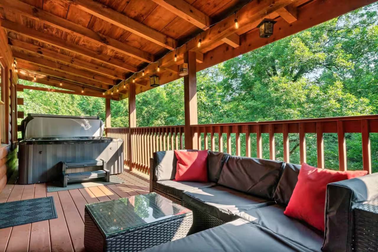 Covered deck with a private hot tub, comfy outdoor seating, string lights, and peaceful wooded views