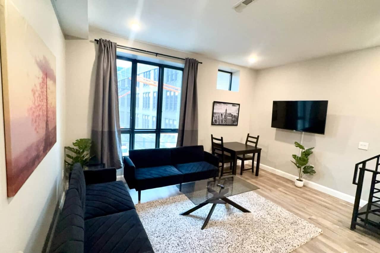 Cozy living room featuring two stylish futon sofas, a flat-screen TV, large windows that fill the space with natural light, and high ceilings that add an airy feel. Perfect for relaxation and entertainment!