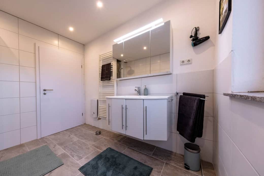 Hell gefliest, mit großem Spiegelschrank einschl. integrierter Beleuchtung sowie einem Unterschrank-Element - das Badezimmer bietet ausreichend Stauraum und Komfort