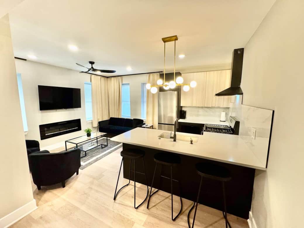 Modern kitchen featuring brand-new appliances, including a breakfast bar, dishwasher, microwave, and refrigerator. It seamlessly connects to the living room, which is furnished with 1sofa bed and 2 sofa chairs, fireplace ,and a flat-screen TV.