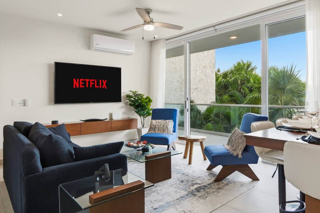 A welcoming living room featuring a comfortable sofa bed, large smart TV, and wide sliding glass doors that open to a private balcony with lush jungle and pool views. A perfect spot to relax, stream your favorites, and enjoy the scenery.