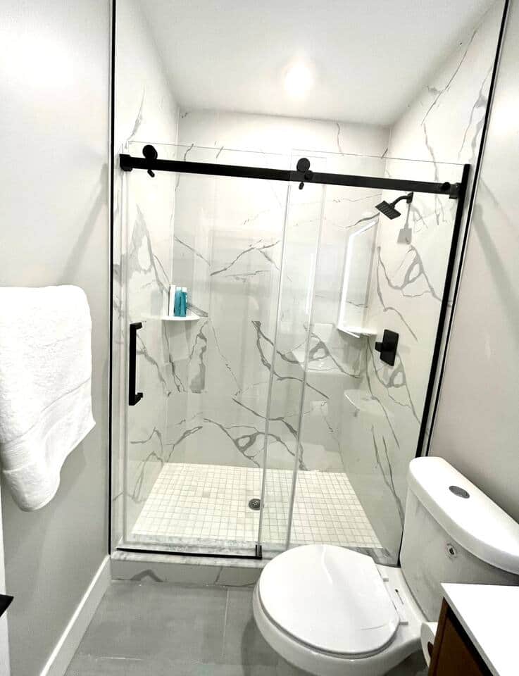 The bathroom boasts a sophisticated carrera tiled shower, with a modern glass door taking center stage. An impressive vanity, complemented by a dazzling lit mirror and sleek black fixtures, adds a touch of sophistication to the overall atmosphere.