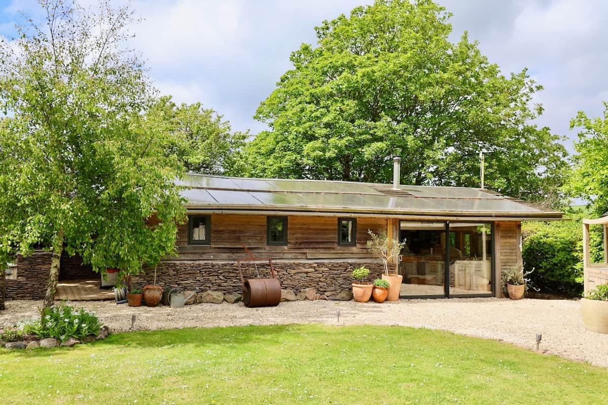 A beautifully converted timber and dry-stone barn with solar panels, sliding glass doors, and a sunny gravel terrace with terracotta planters  set in lush, tree-lined countryside.