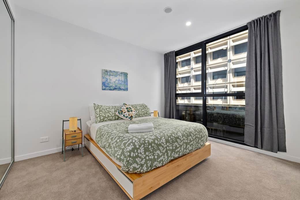 This inviting bedroom features a large window that lets in abundant natural light, creating a serene atmosphere. The bed is dressed in a fresh, floral comforter. The stylish bedside table and tasteful wall art, makes this room a perfect retreat.