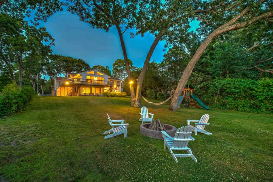 Fire pit with Adirondack chairs on the lawn at dusk, with the house lit up behind
