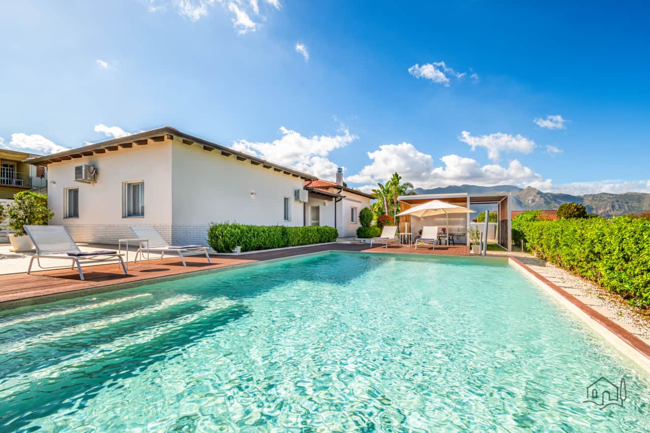 Crystal-clear pool with the villa facade, a shaded dining area, and breathtaking mountain views.
