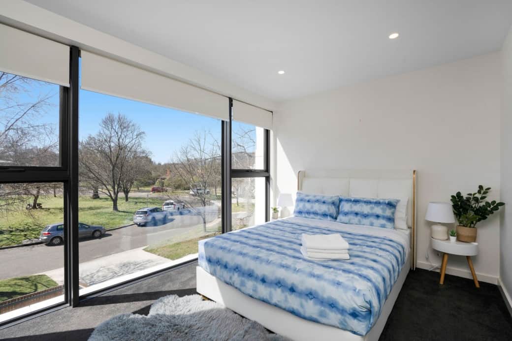 Spacious bedroom with floor-to-ceiling windows offering stunning views of the trees and street. The room features a large, comfortable bed with soft blue bedding and fresh towels. Natural light fills the space, complemented by modern furnishings.