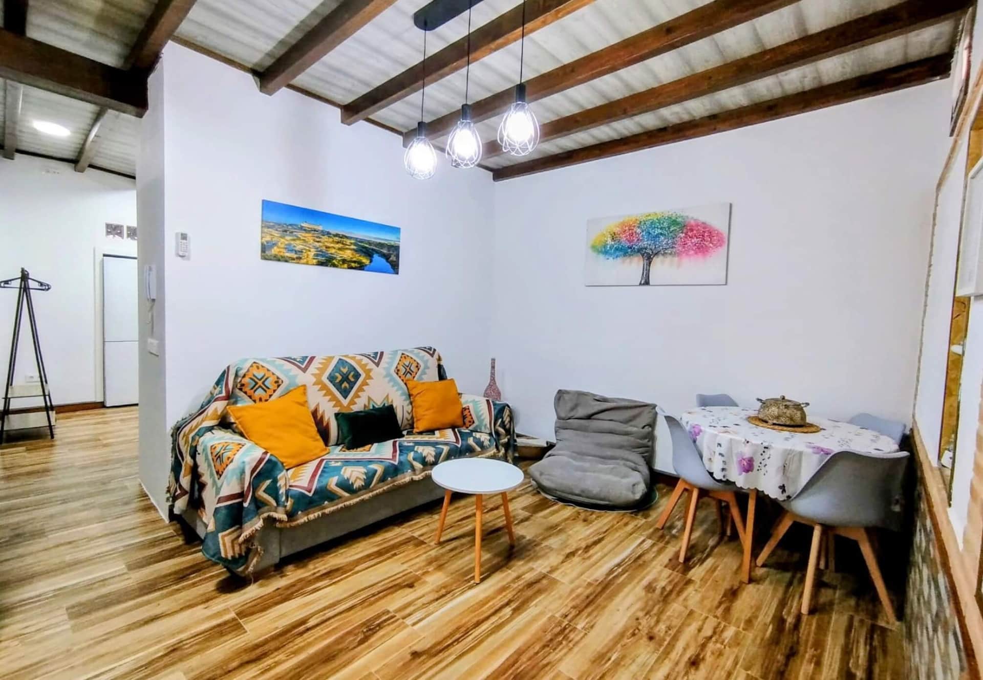 Salón acogedor con sofá gris decorado con manta de colores y cojines mostaza, mesa de centro blanca, puff gris y comedor con mesa redonda