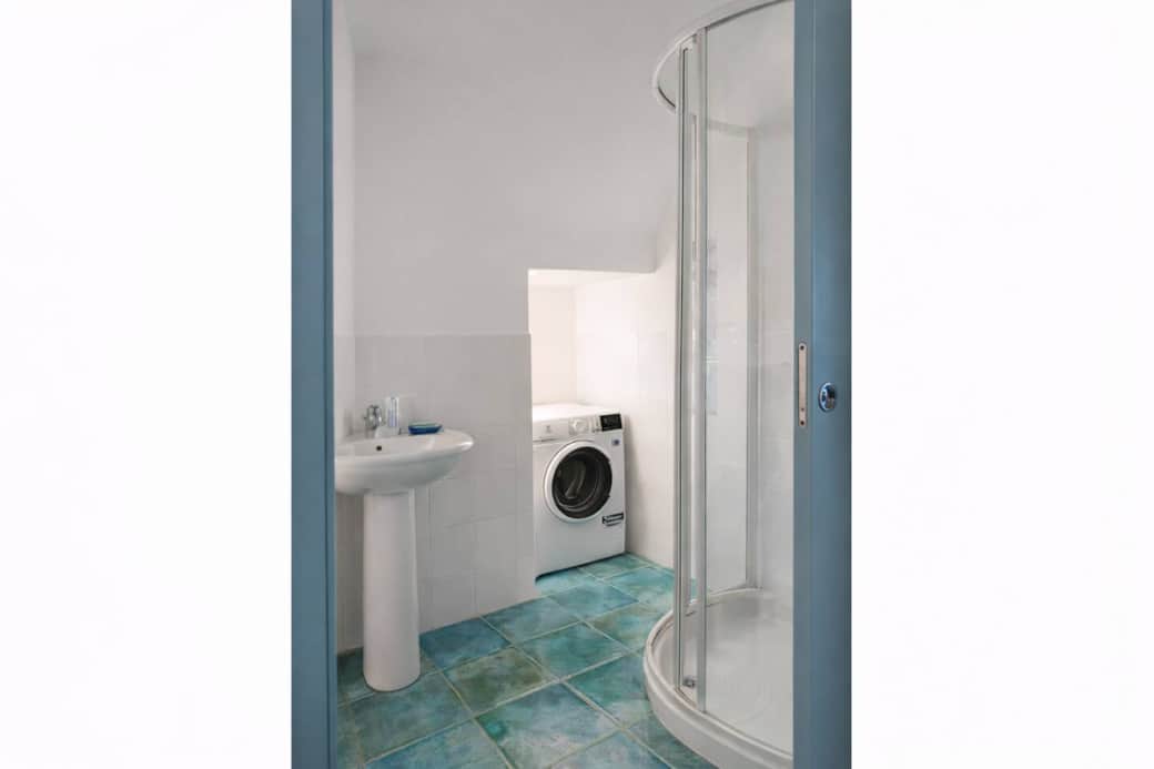 Bagno luminoso e funzionale con ampia doccia angolare, lavabo con specchio e dettagli in tonalità turchese che richiamano il pavimento. Completo di lavatrice, ideale anche per soggiorni più lunghi, offre uno spazio pratico e ben organizzato.