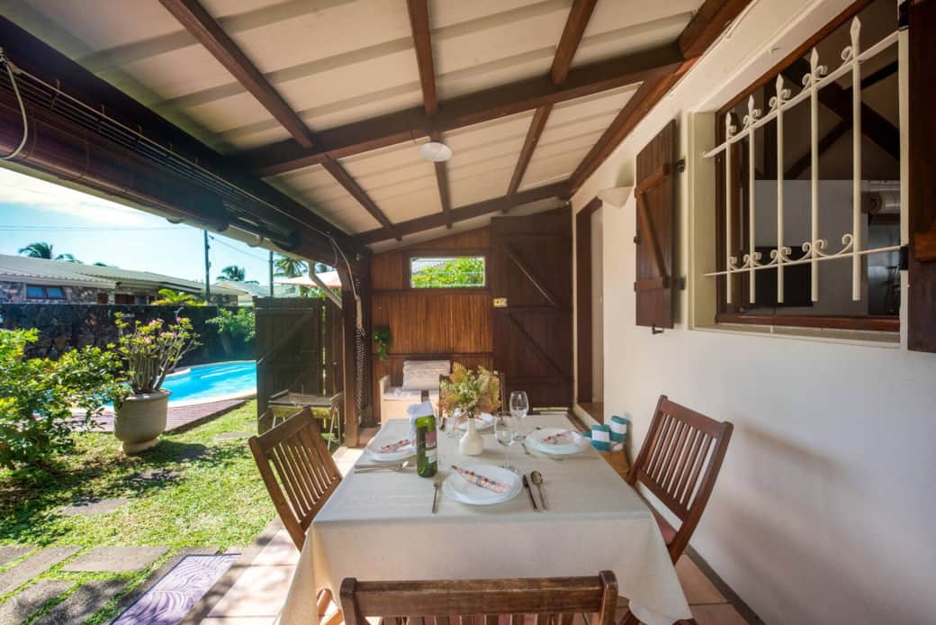 Charming covered terrace with a dining table set for four, overlooking a lush garden and shared pool
