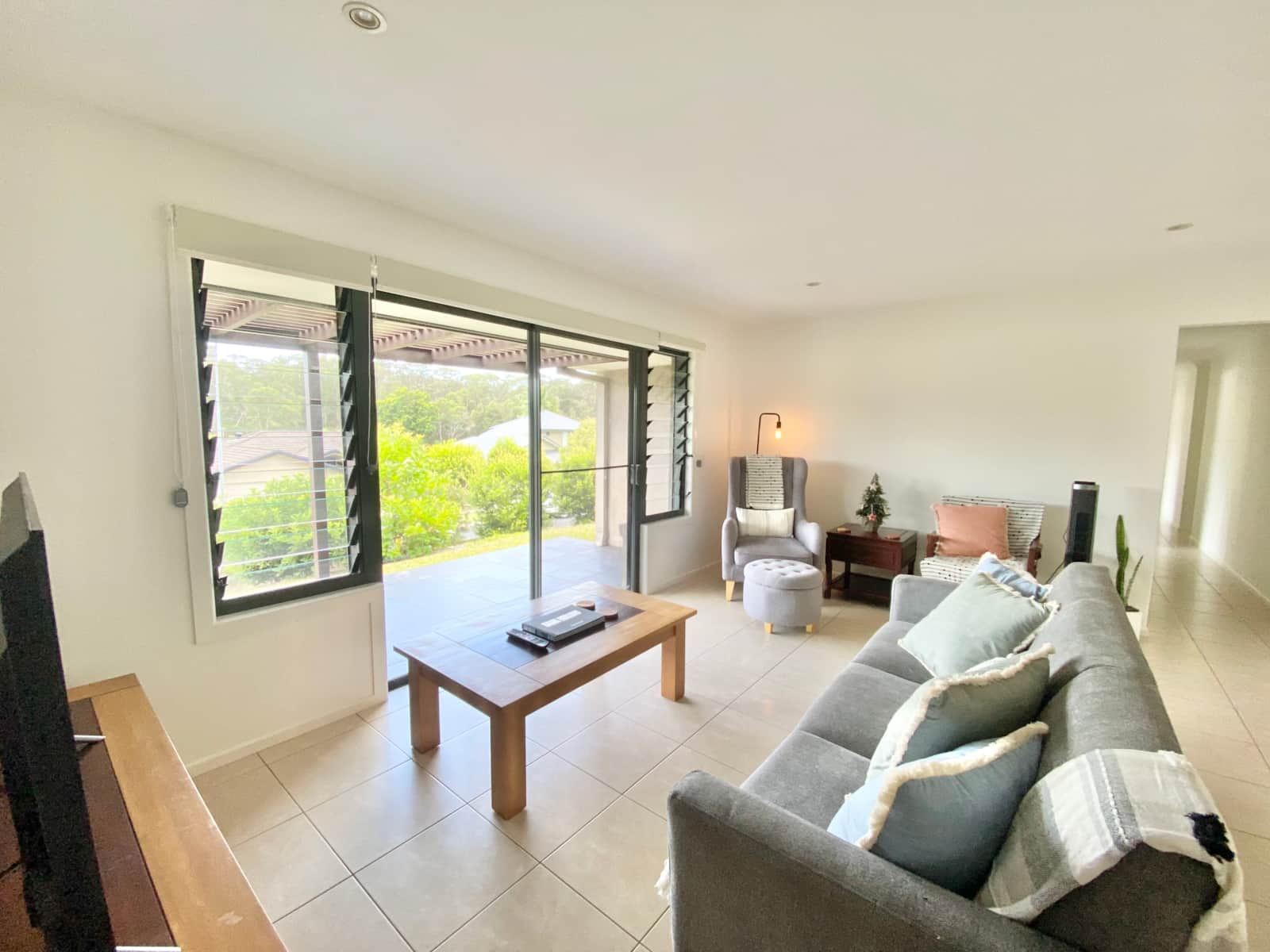 Bright open-plan living room with grey sofas, armchair and leafy views through the sliding doors at The Lovely, Boambee East