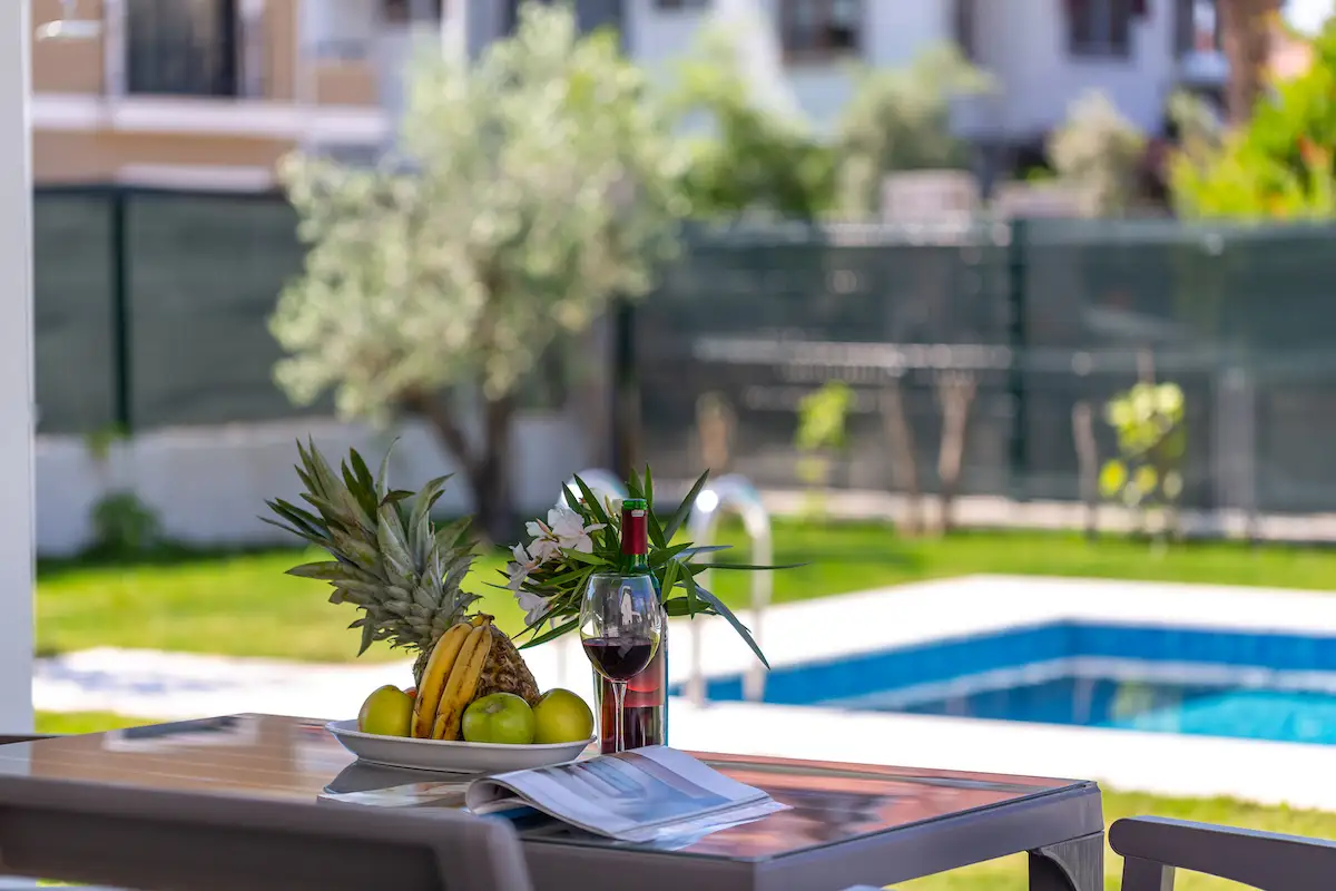 Table set with fruit, a bottle of wine, and a glass, with a pool and garden in the background at Villa Nergis 1.