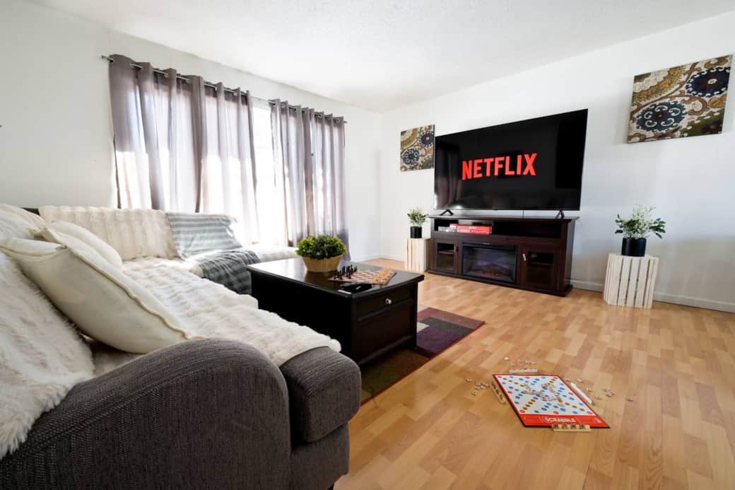 A bright living room with large windows, a cozy sectional sofa with soft throws, and a stylish coffee table with a chess set. Enjoy the natural light, wooden floors, and a peaceful space to relax.