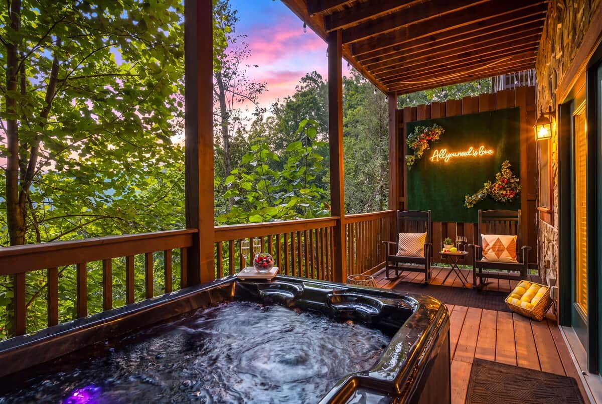 Romantic mountain‑view deck with a bubbling hot tub, cozy rocking chairs, and a glowing ‘All You Need Is Love’ neon wall—creating the perfect space for couples to unwind together as the sunset colors the sky.
