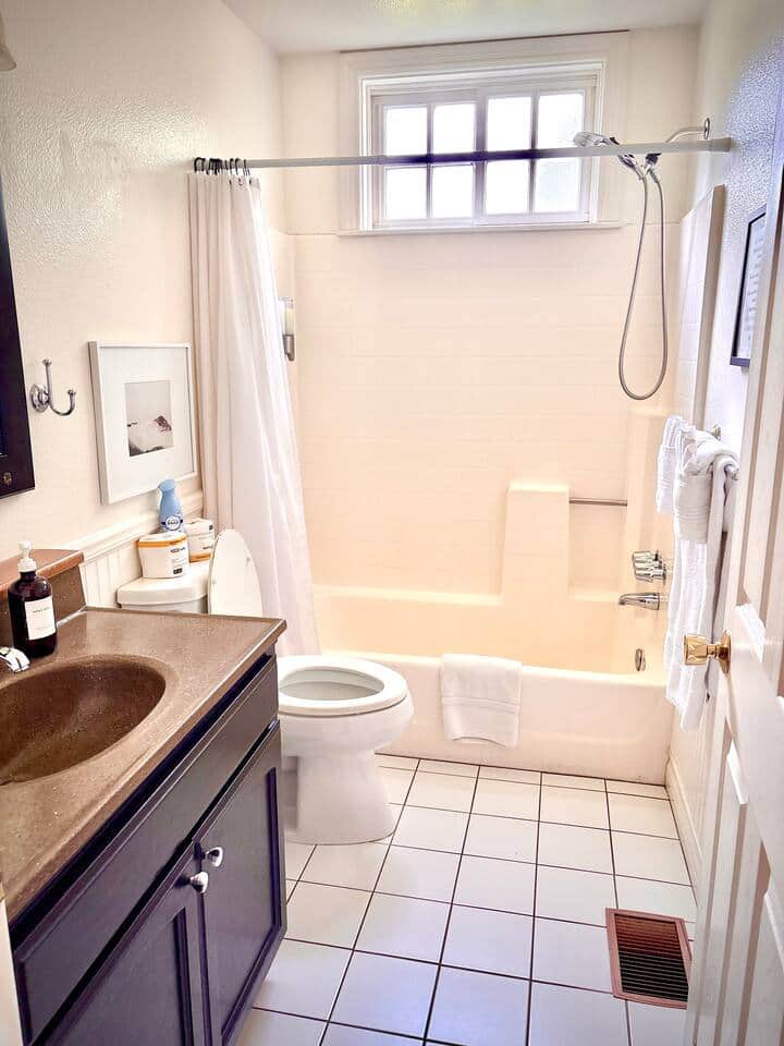 Main floor hallway full bathroom with tub/shower combo.  Vanity with framed mirror, white fixtures, and high window for natural light while maintaining privacy. Fresh towels and essential amenities provided.