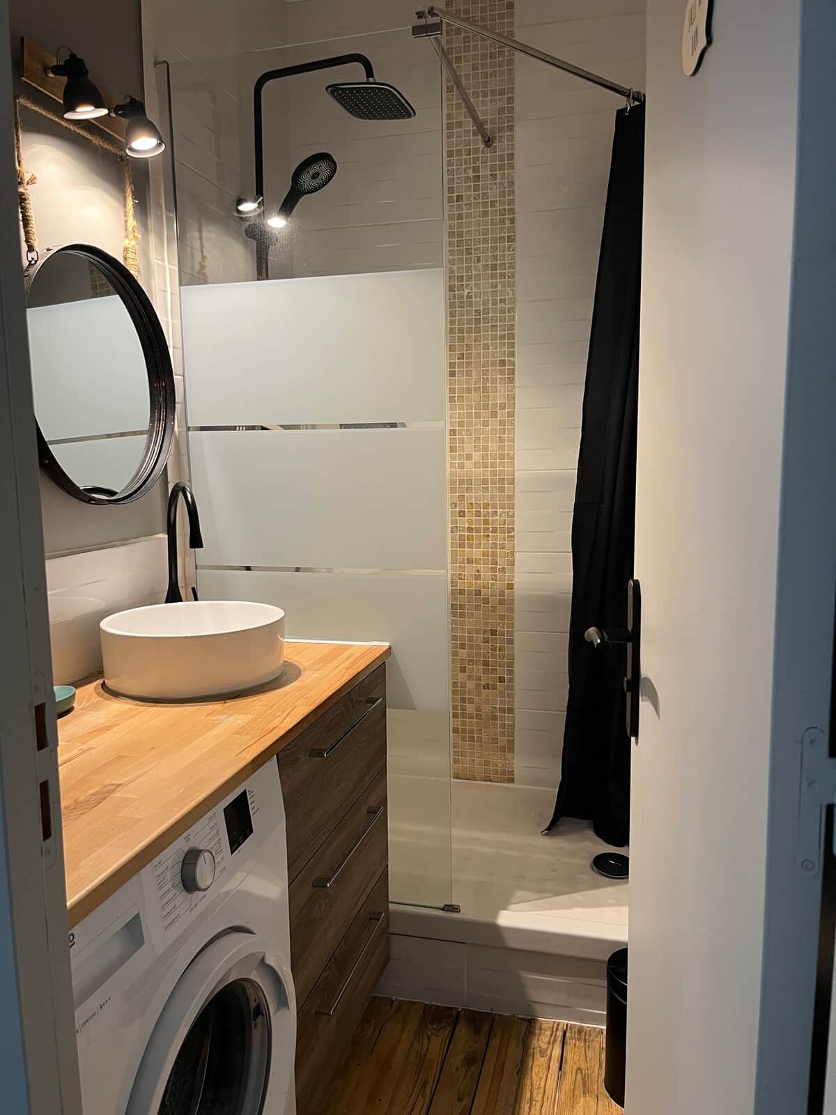 Salle de bain moderne avec douche, paroi vitrée et mosaïque beige/blanche. Meuble bois avec vasque ronde, miroir suspendu, machine à laver intégrée et éclairage chaleureux.