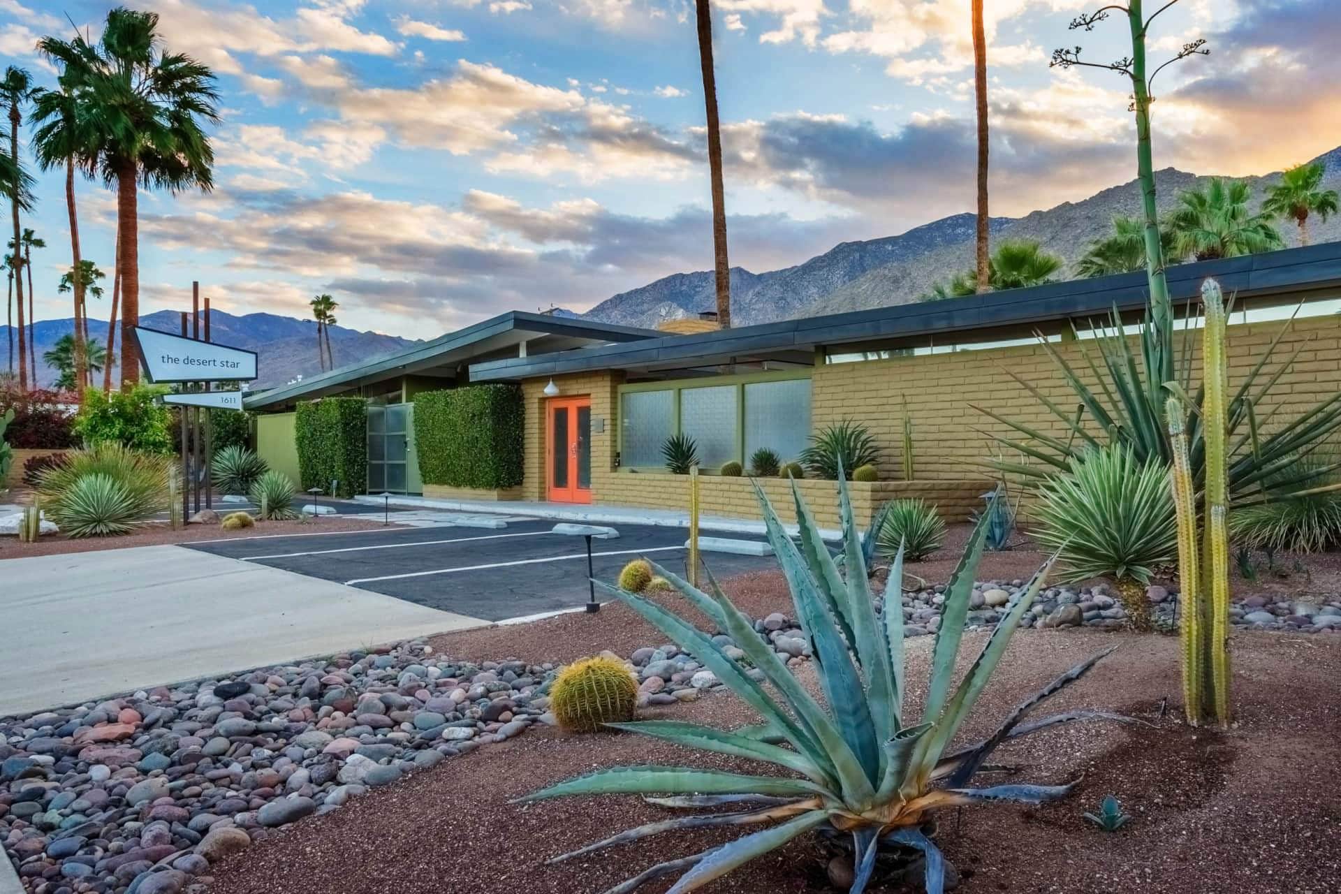 A single-story building with a flat roof and large windows, surrounded by desert landscaping and palm trees, with a mountain range in the background.