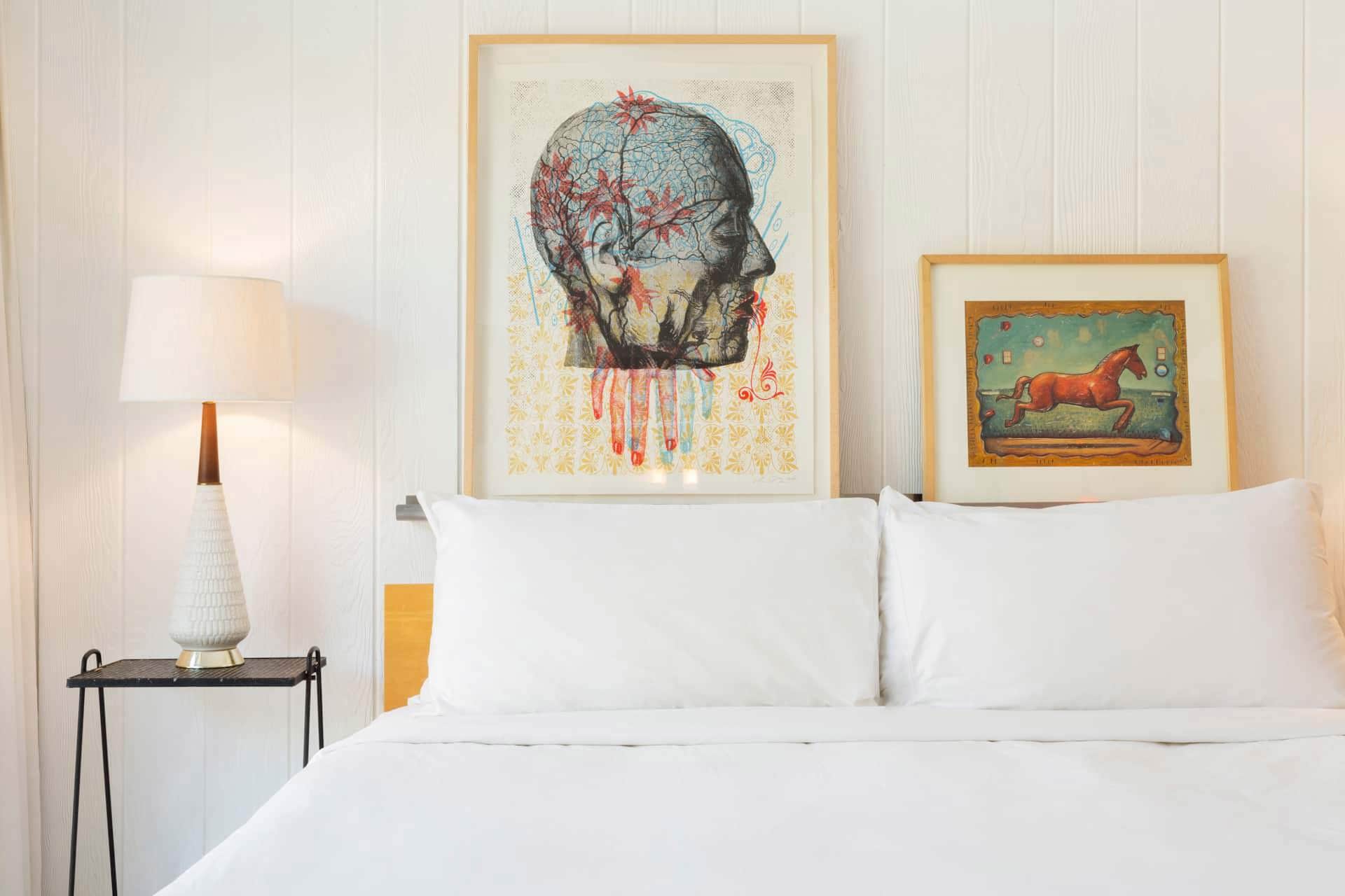 A bed with white linens and two pieces of artwork above it