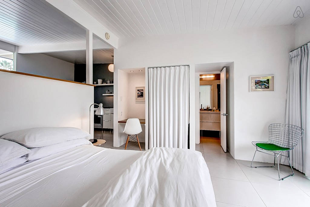 A wide view of a white suite with a bed, desk, chair, and doorway to another room.