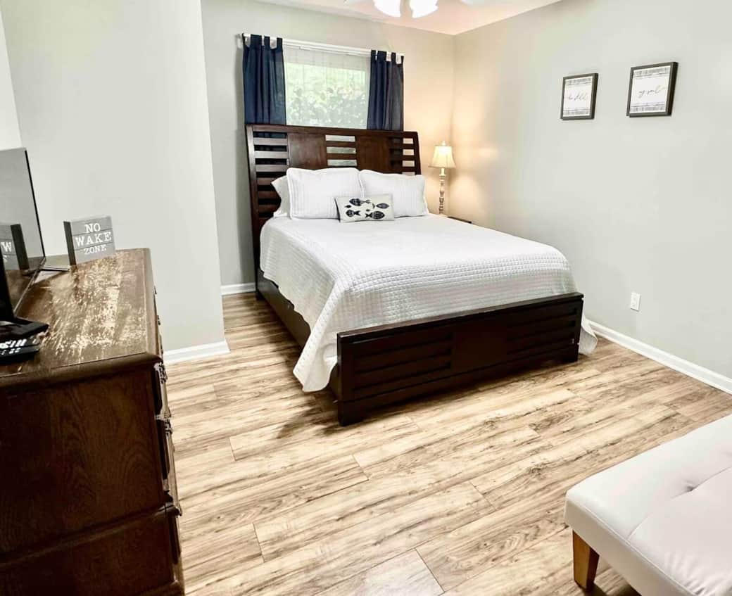 Sun-drenched queen guest room with honey-wood bed, navy curtains, and extra sleeper seating. Queen bed, lake-themed decor, flat-screen TV, gray walls, coastal accent pillow, driftwood flooring, Smith Mountain Lake getaway.