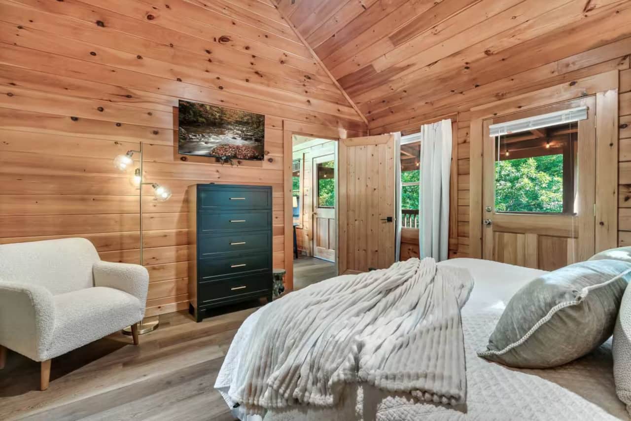 Cozy wood-paneled bedroom with vaulted ceilings, a plush bed, reading chair, and access to deck